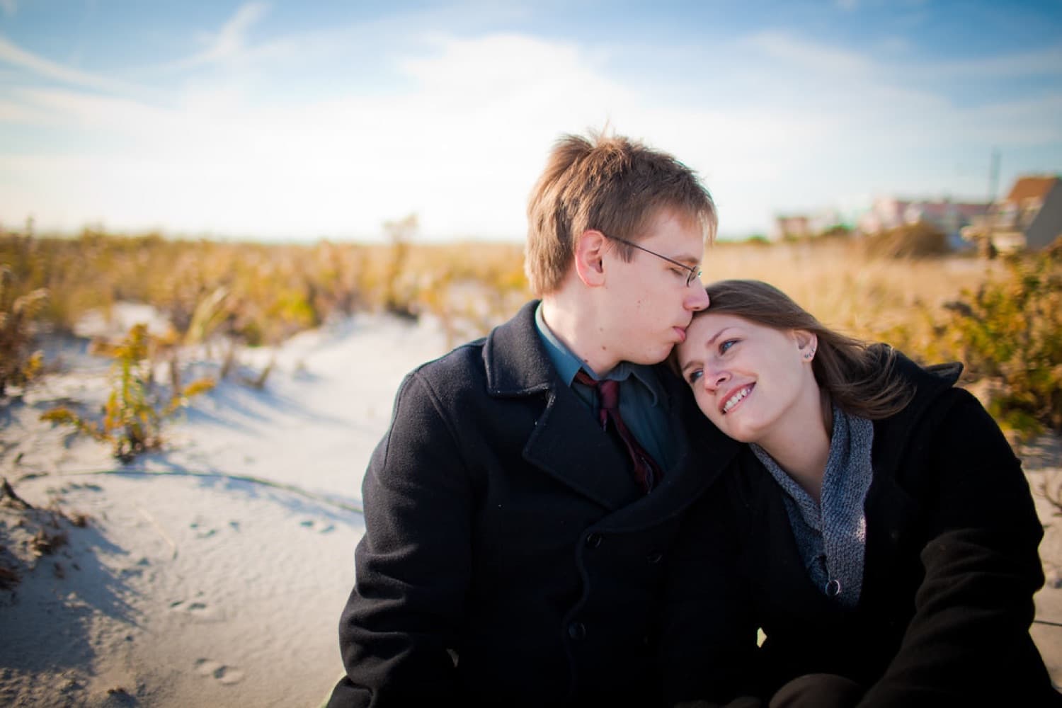 Engaged couple close together in winter coats by the dunes